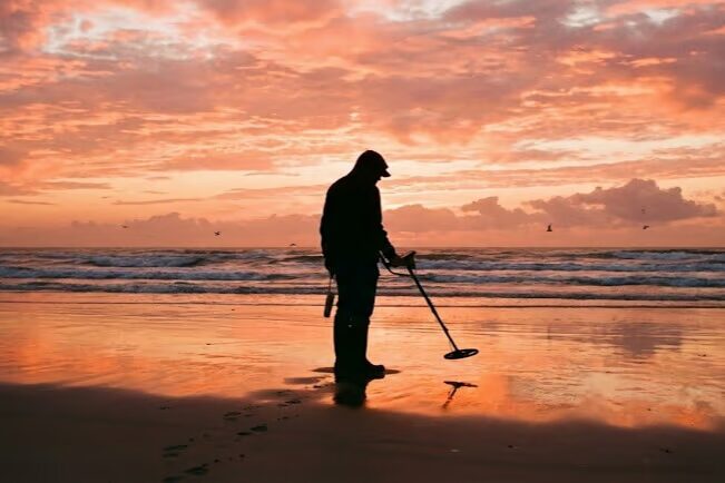 Metal detectorist using detector on sandy beach at sunrise hunting for jewelry and coins near ocean waves