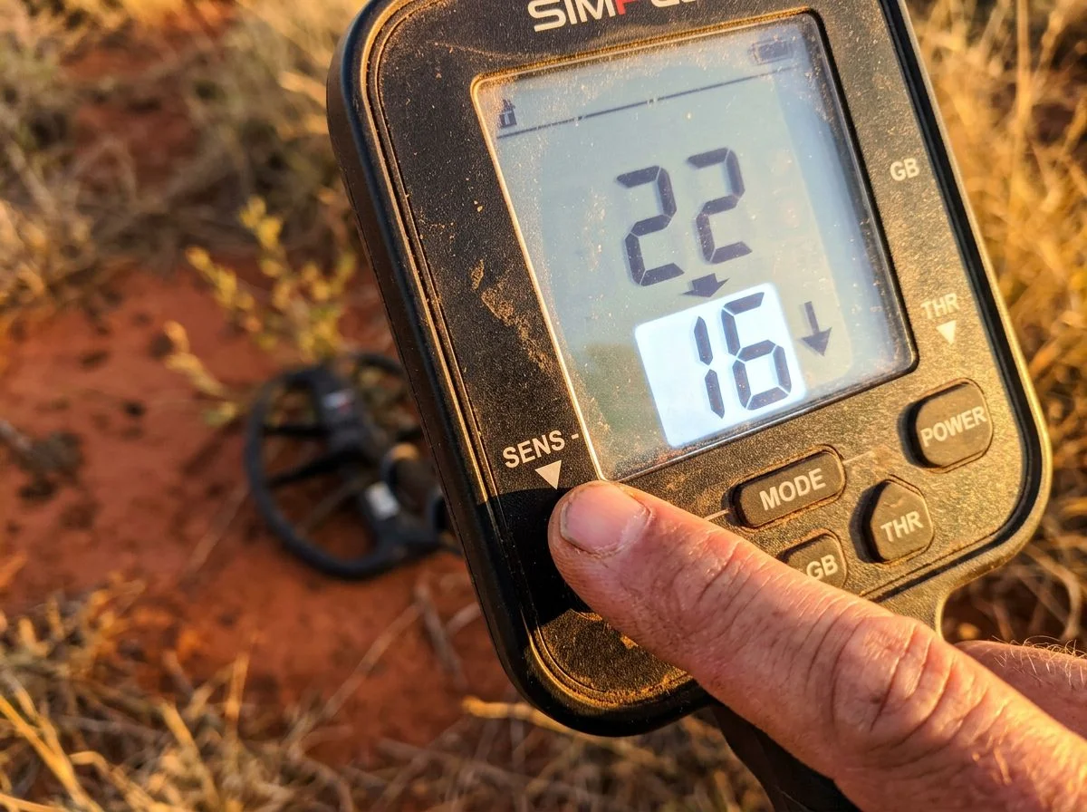 Finger pressing sensitivity button on metal detector control panel, reducing level from 22 to 16 over red clay ground